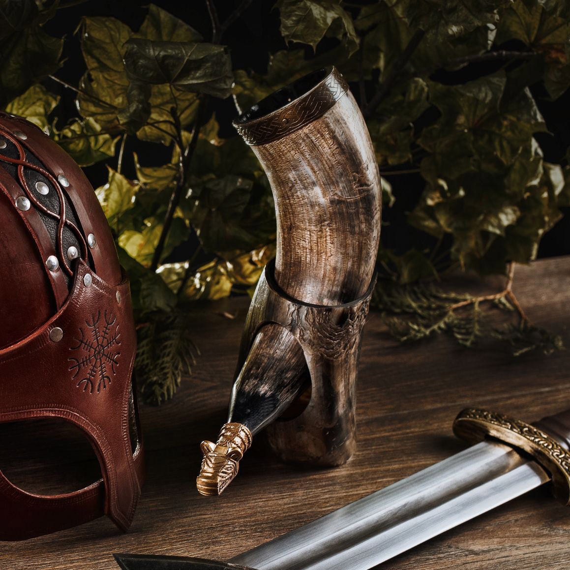 Decorative horn, leather helmet, and sword on a wooden surface with green foliage in the background