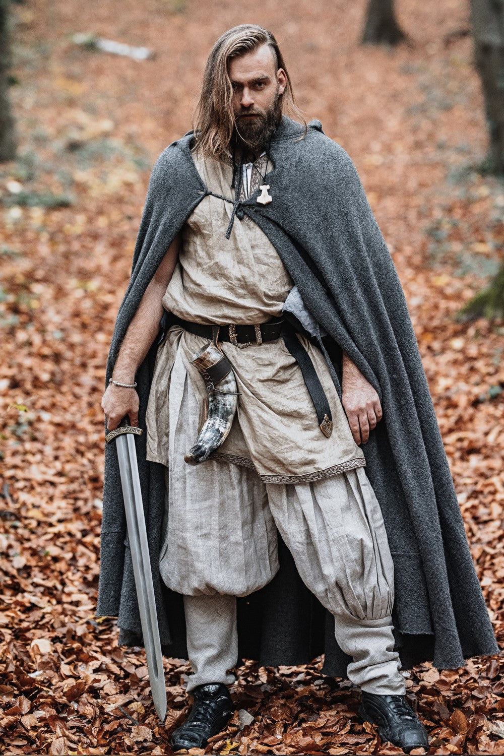 Man in historical warrior attire with a sword standing in a forest.