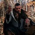 Man in medieval attire holding a large tusk and a helmet in a forest setting