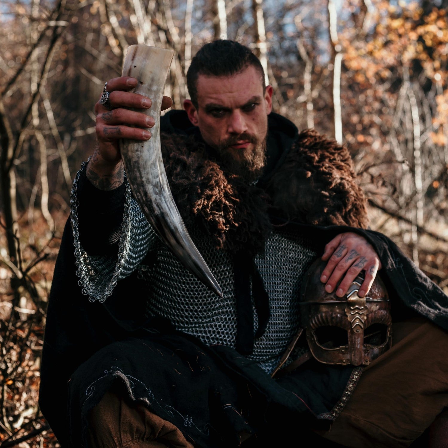 Man in medieval attire holding a large tusk and a helmet in a forest setting