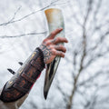 Hand holding a large drinking horn with intricate designs, against a blurred natural background.