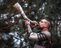 Man in warrior costume blowing a large metal horn against a forest background