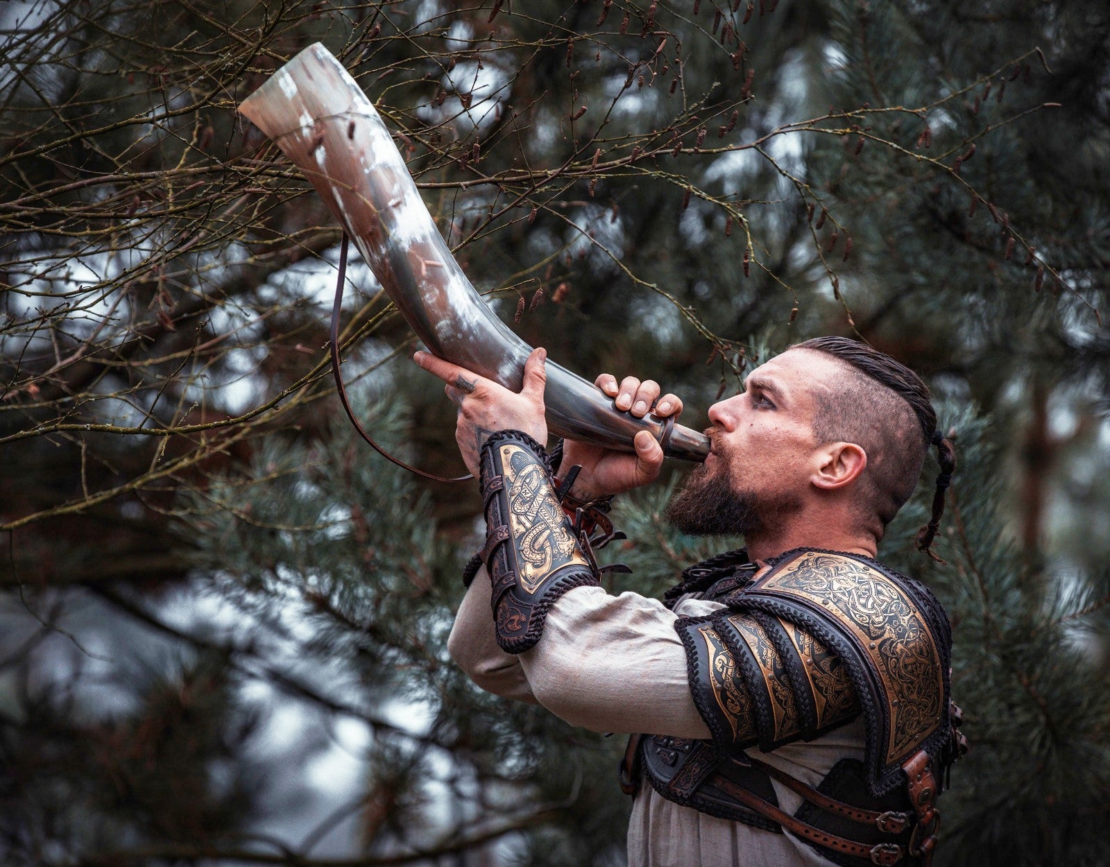 Man in warrior costume blowing a large metal horn against a forest background