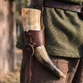 Tusk in a leather holder on a person wearing green fabric and brown leather belt outdoors.