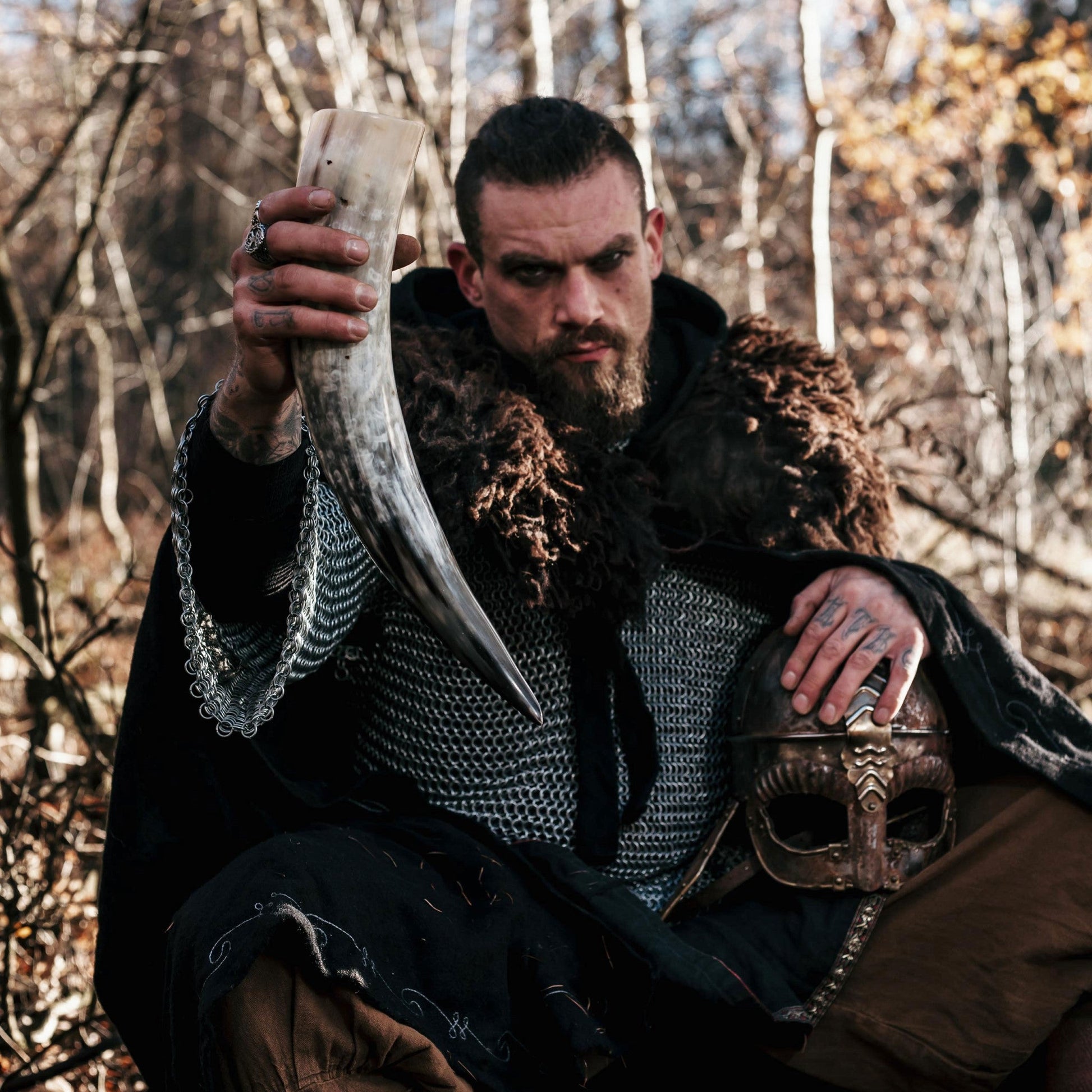 Man in medieval attire holding a large tusk and a helmet in a forest setting