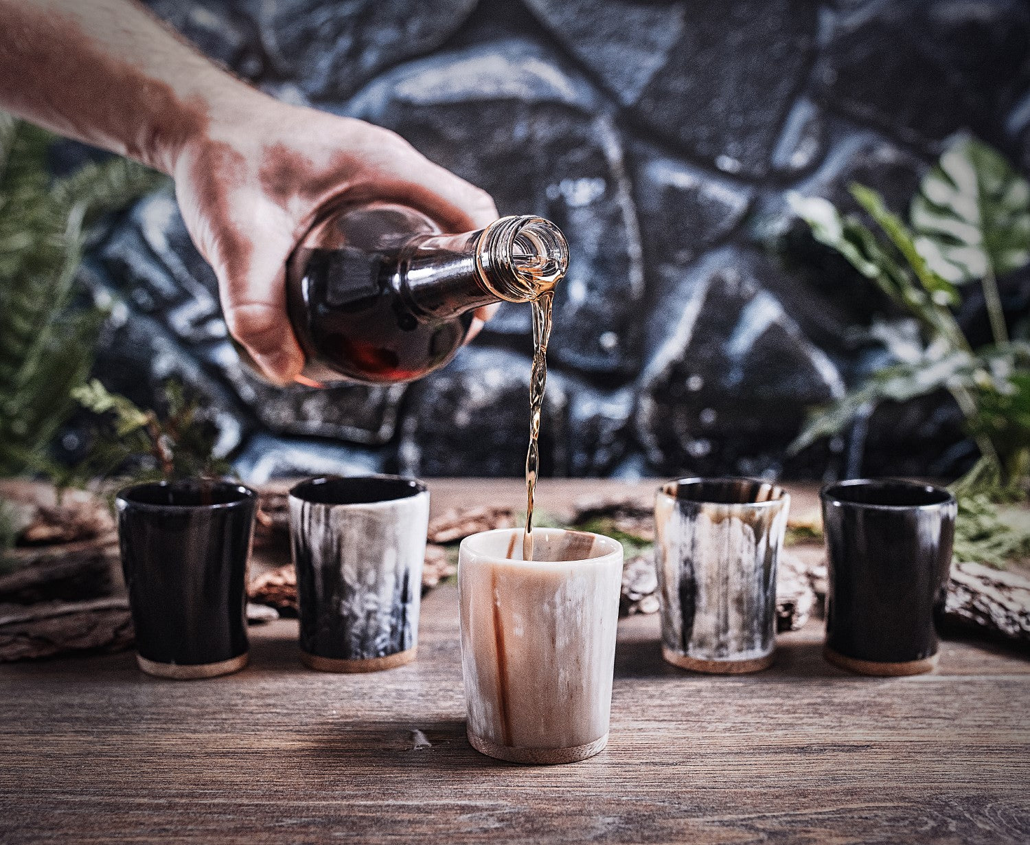 Hand pouring liquid from a bottle into a wooden cup on a rustic wooden table with other cups.