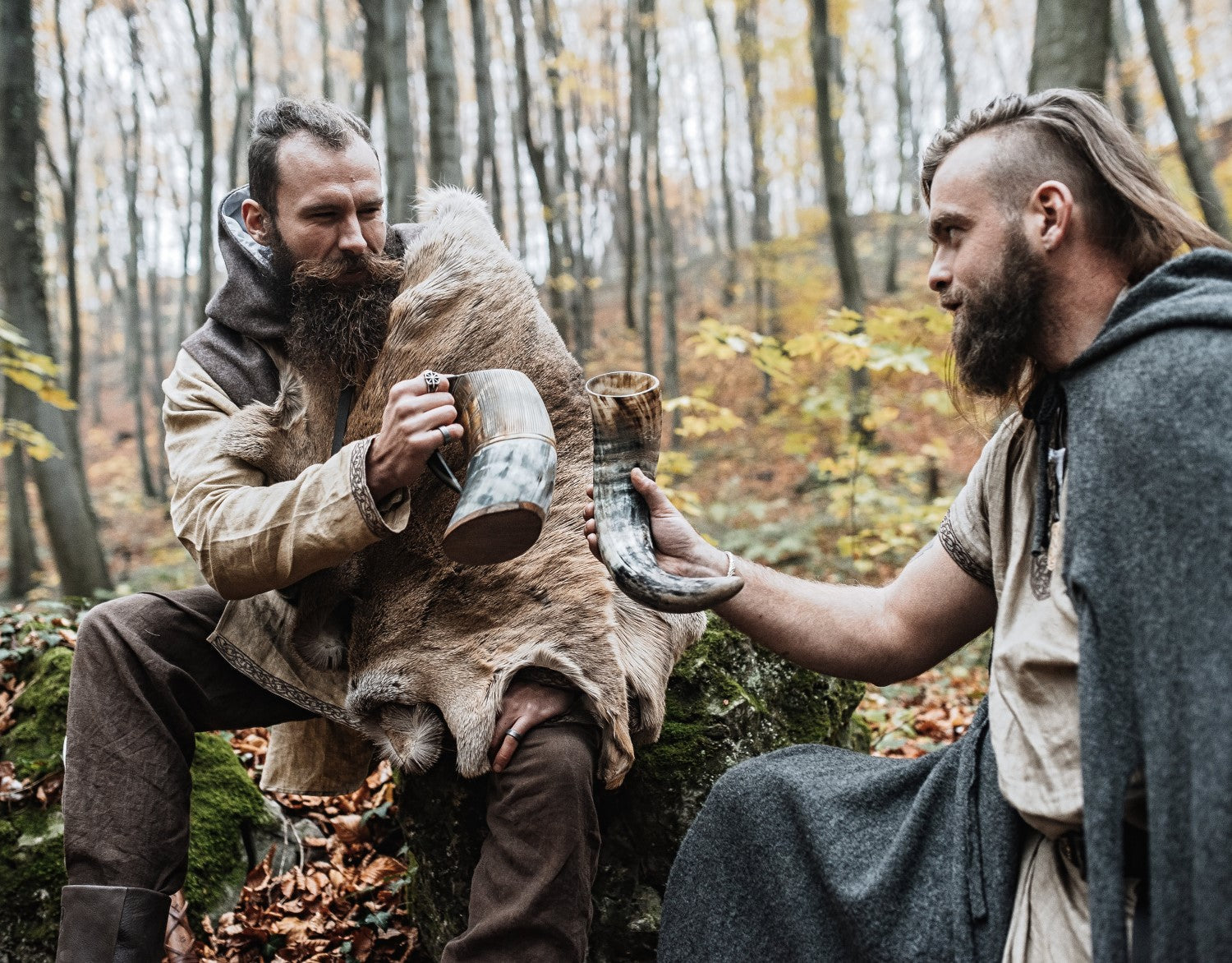 Two men in medieval attire sitting in a forest with a deer head.
