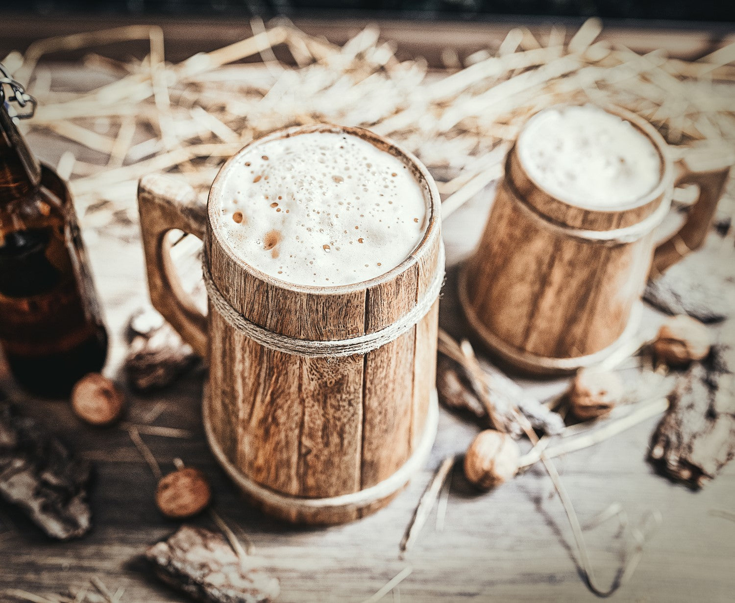Two wooden mugs with a frothy beverage on a rustic surface with nuts and a bottle.