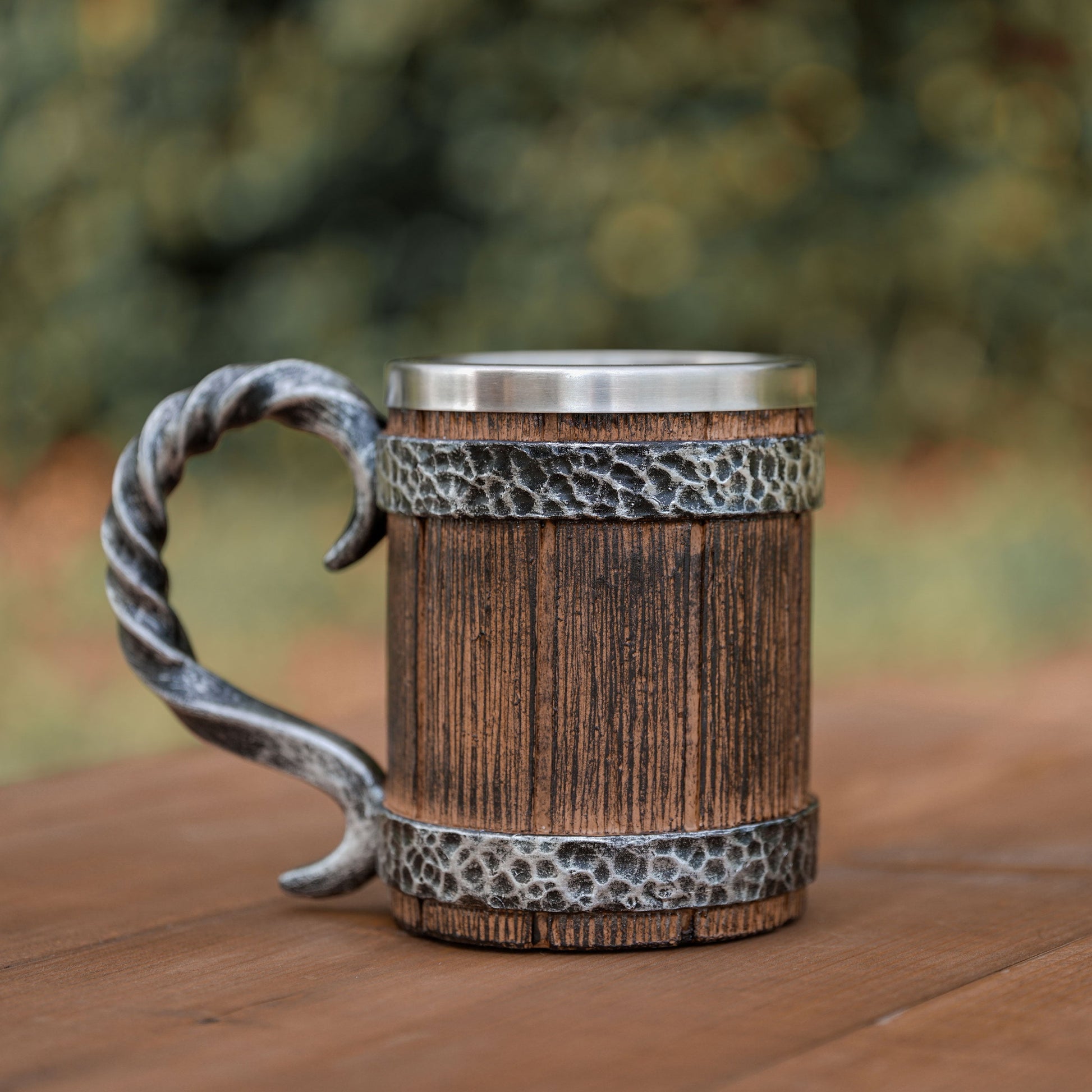 Wooden mug with metal handle and band on a wooden surface with blurred greenery in the background