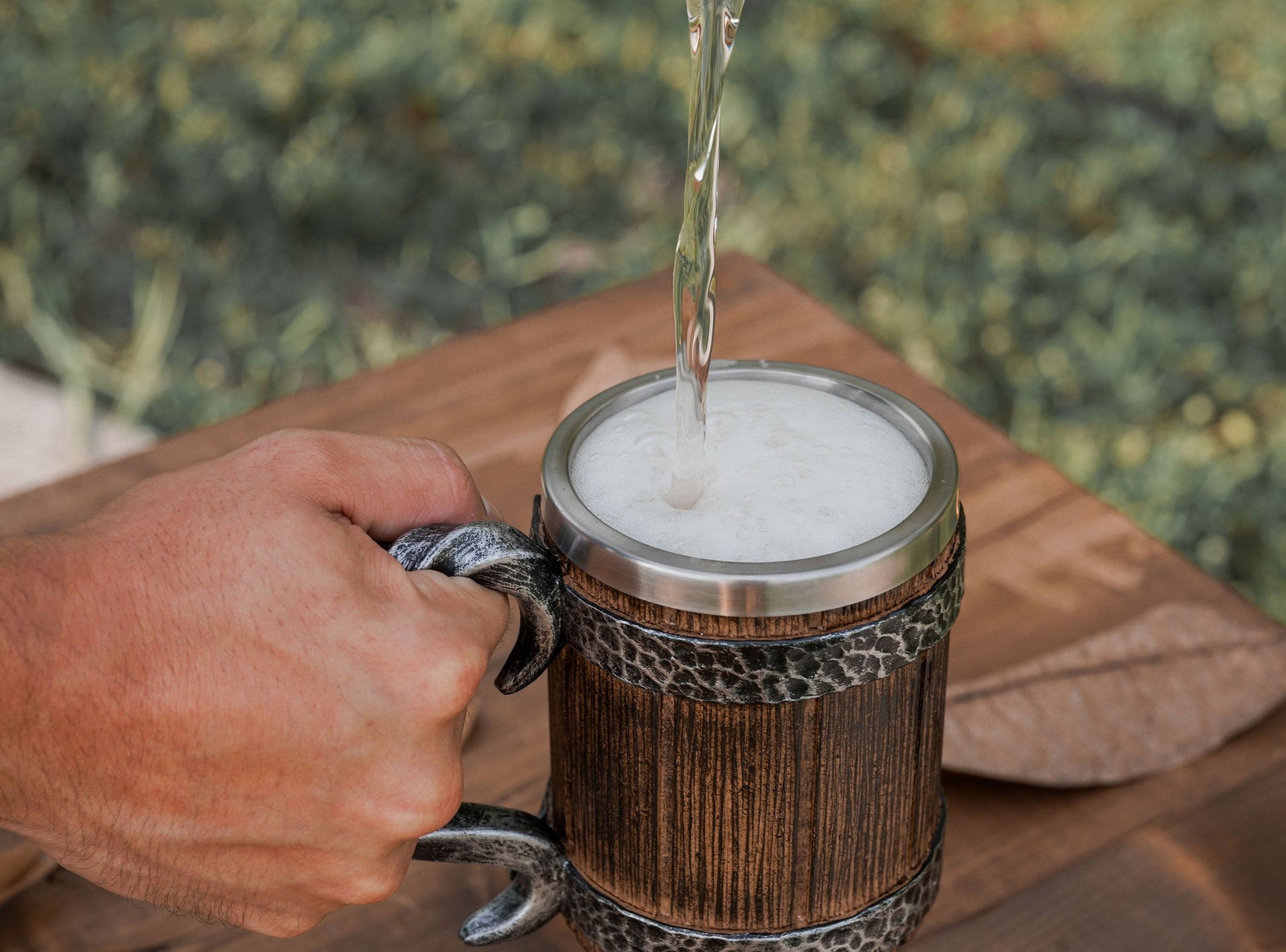Hand pouring liquid into a wooden mug with a metal lid on a wooden surface.